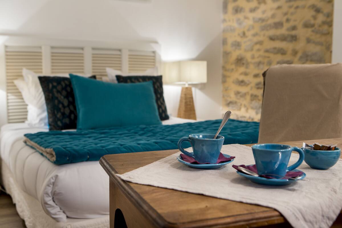 A neatly made bed with teal bedding, a wooden nightstand with two blue cups and saucers, a lamp, and a stone-patterned wall.
