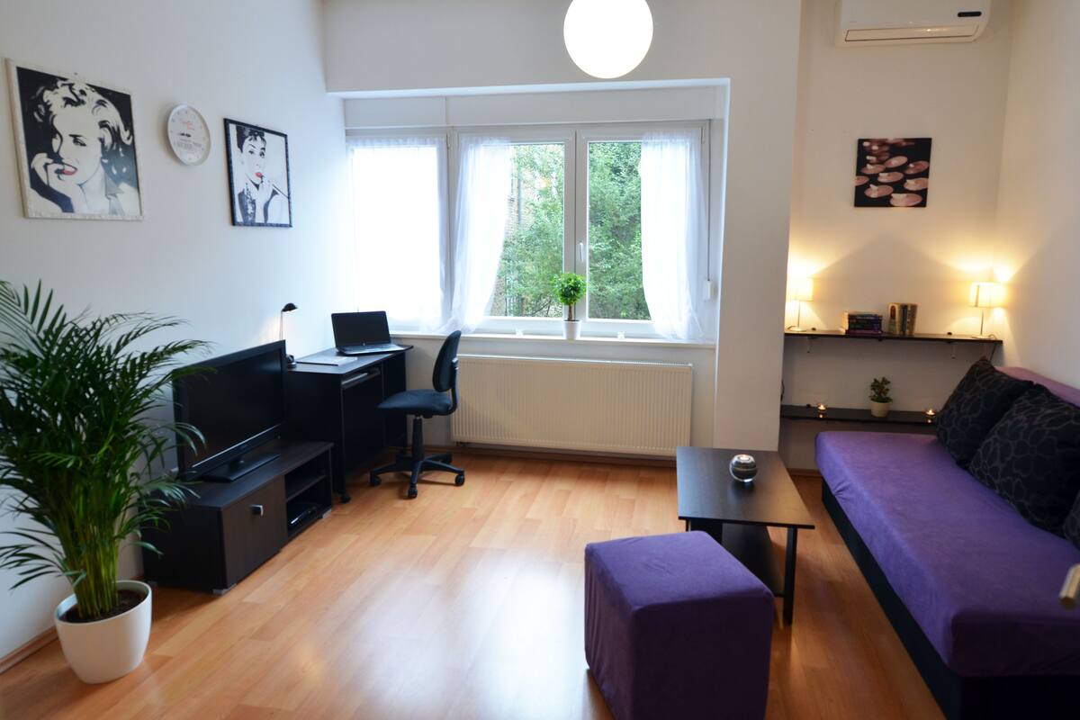 A modern living room with a black desk, purple sofa, and a large window with curtains.