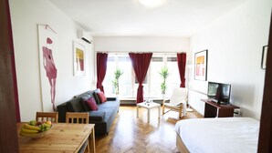 A modern hotel room with a wooden floor, a bed, a sofa, a dining table with fruit, a TV, and two potted plants. - First Choice Centar Apartments (Zagreb)