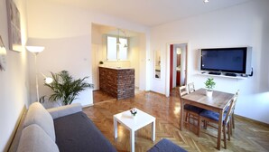 A modern living room with a sofa, a coffee table, a dining table with chairs, a TV mounted on the wall, and a brick fireplace. - First Choice Centar Apartments (Zagreb)