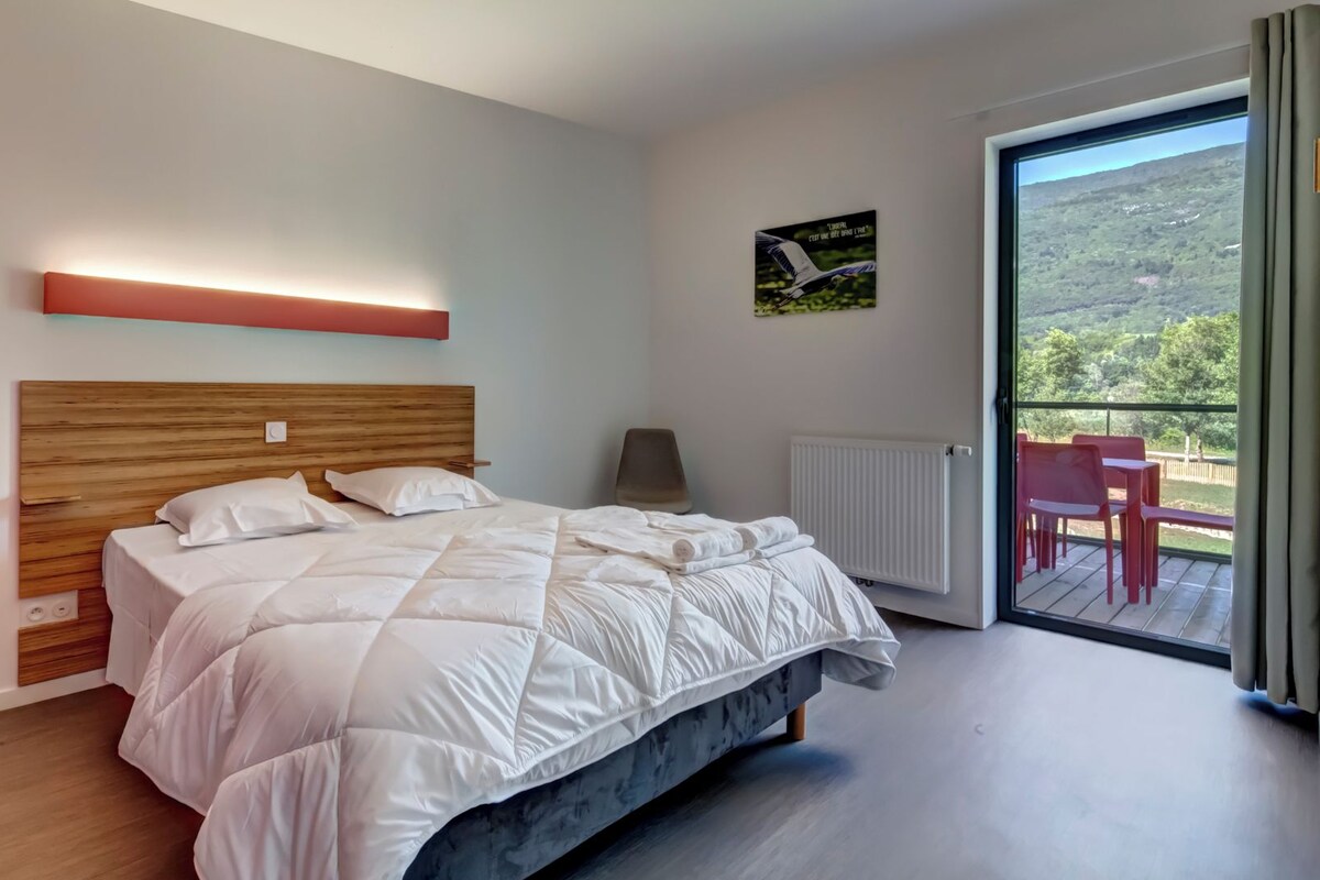 Une chambre moderne avec un grand lit, une table de chevet, une chaise et une vue sur un balcon aménagé avec des sièges d’extérieur.