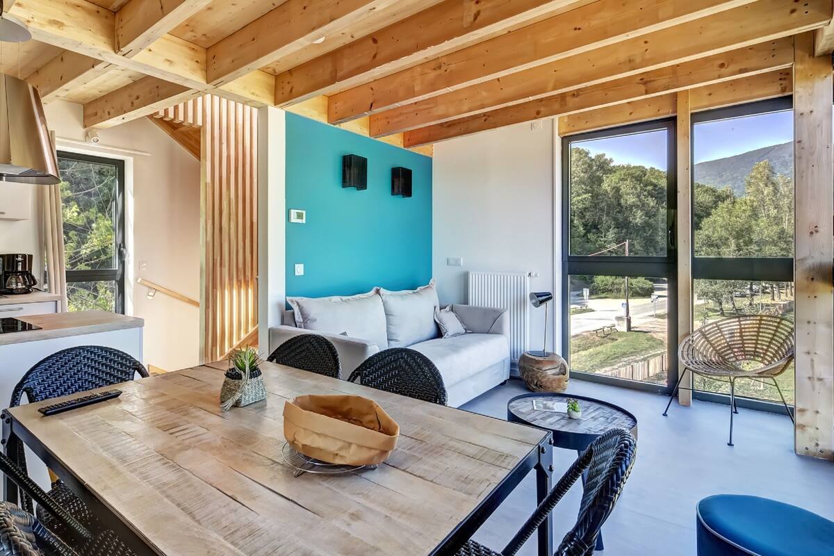 Ein modernes Interieur mit Holzboden, einer Essecke mit Tisch und Stühlen, einer Couch und einem großen Fenster mit Blick ins Freie.