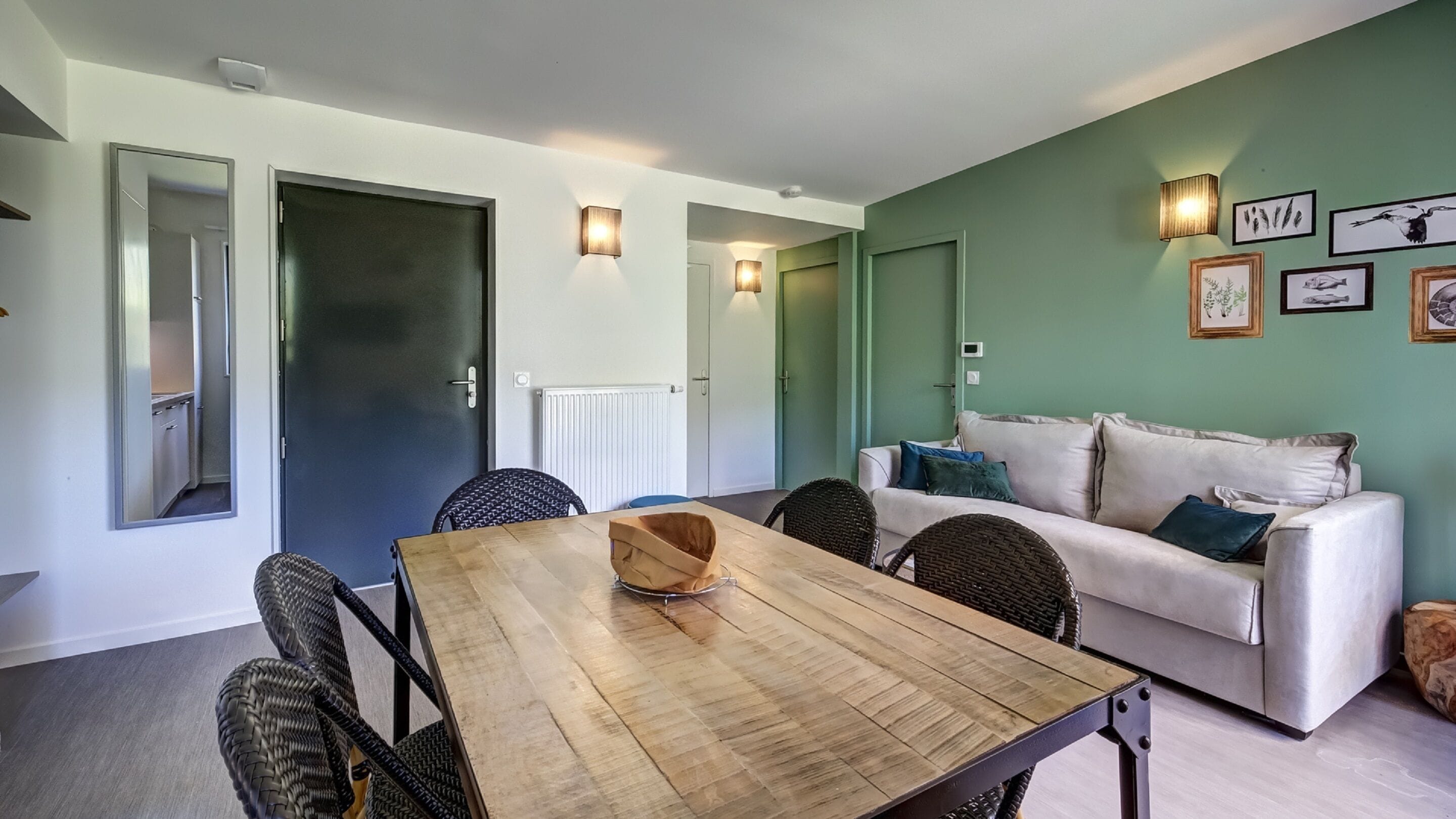 A modern living room with a wooden dining table, wicker chairs, a white sofa with cushions, and wall-mounted lights.