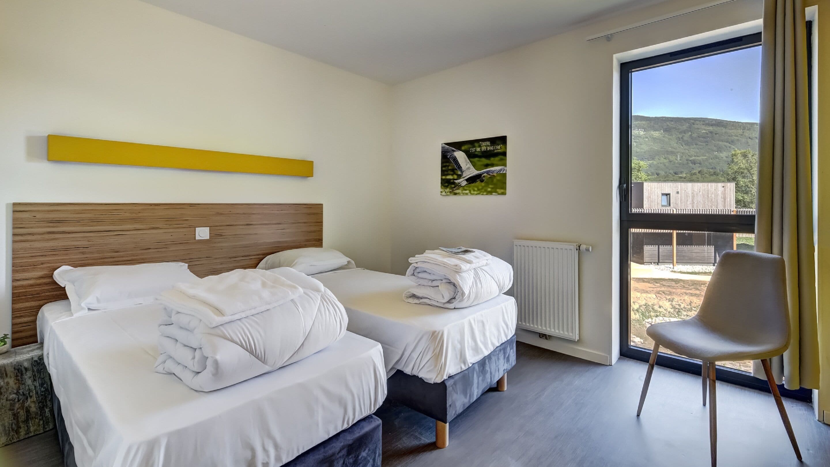 Une chambre d’hôtel moderne avec deux lits, une grande fenêtre donnant sur la campagne et un fauteuil près de la fenêtre.