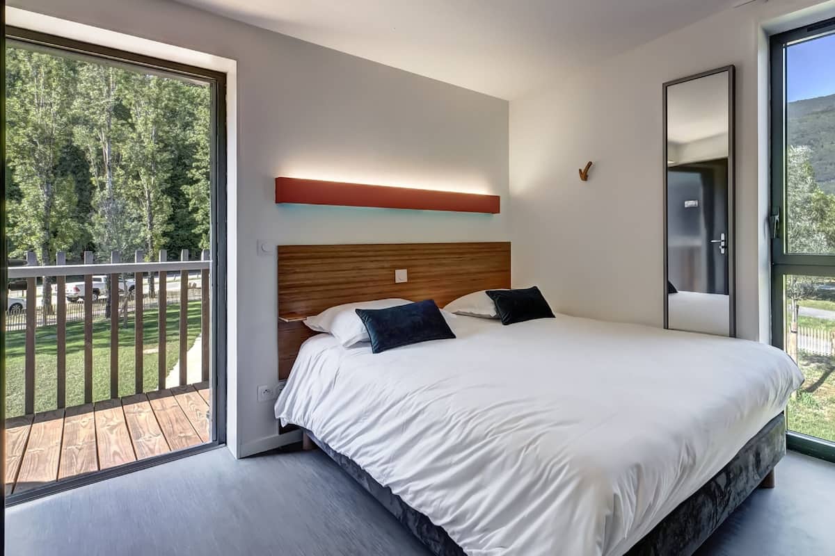 Ein modernes Hotelzimmer mit einem großen Bett, einem hölzernen Kopfteil, einem Balkon mit Blick auf Bäume und Hügel und einem großen Fenster mit Vorhängen.