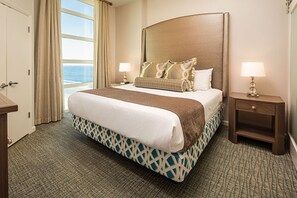 A hotel room with a large bed, two bedside tables, a window with curtains, and a view of the ocean.