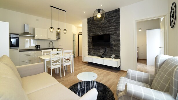 A modern living room with a sofa, armchair, dining table, and chairs. There is a kitchen area with cabinets and a stove. A fireplace with a TV is visible, and a clock is on the wall. - First Choice Centar - Primorska (Zagreb)