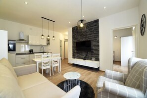A modern living room with a sofa, armchair, dining table, and chairs. There is a kitchen area with cabinets and a stove. A fireplace with a TV is visible, and a clock is on the wall. - First Choice Centar - Primorska (Zagreb)