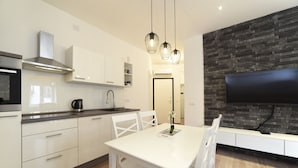 A modern kitchen with white cabinetry, a stainless steel range hood, a built-in oven, and a black countertop. There is a dining table with white chairs, a vase with flowers, and a flat-screen TV mounted on a stone-textured wall. - First Choice Centar - Primorska (Zagreb)