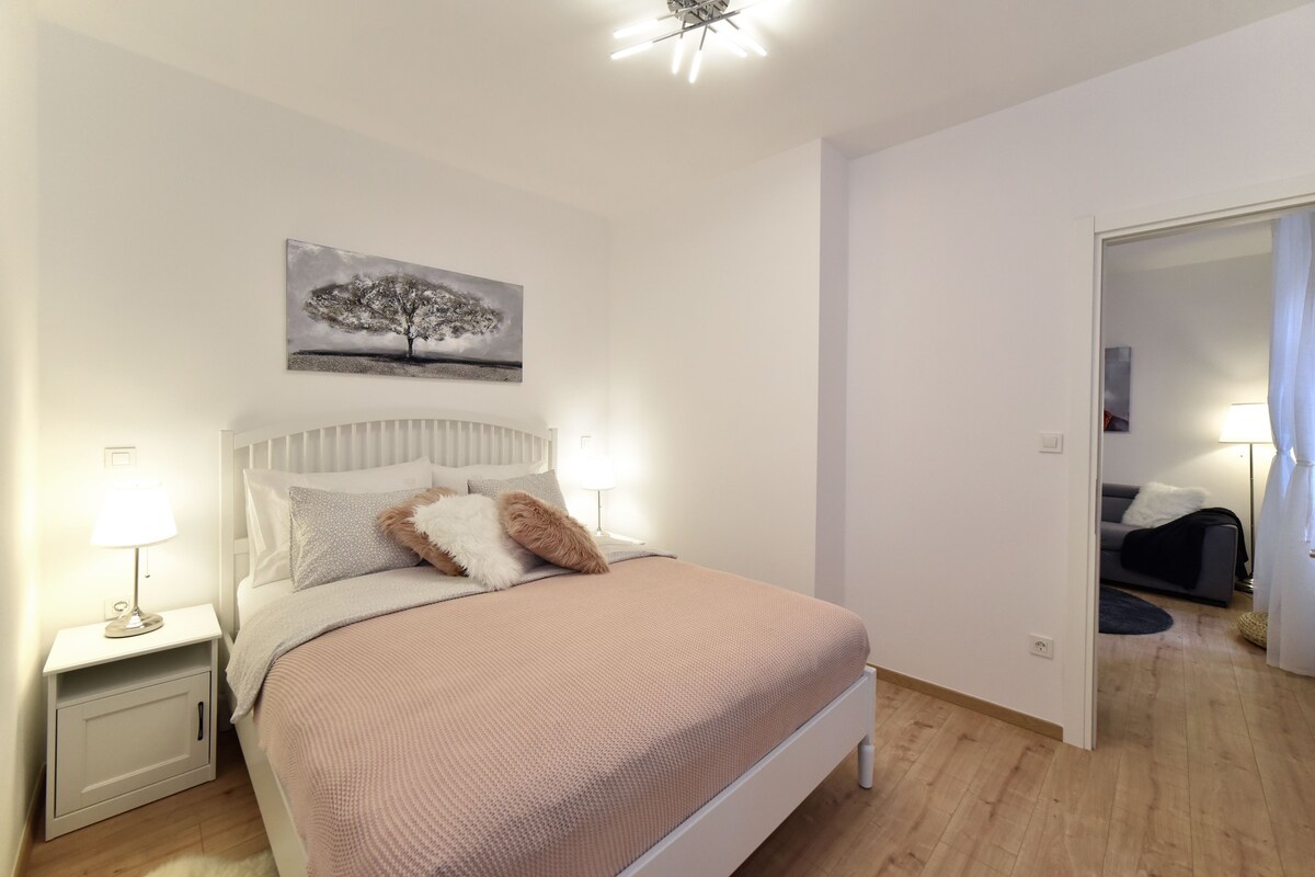 A bedroom with a bed, nightstands, a lamp, a painting, and a door leading to another room.