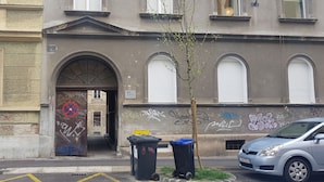 A building with a large arched entrance, graffiti on the walls, and two trash bins on the sidewalk. - First Choice Centar - Primorska (Zagreb)