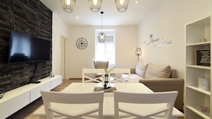 A modern living room with a flat-screen TV, a stone accent wall, a dining area with white furniture, and decorative wall art. - First Choice Centar - Primorska (Zagreb)