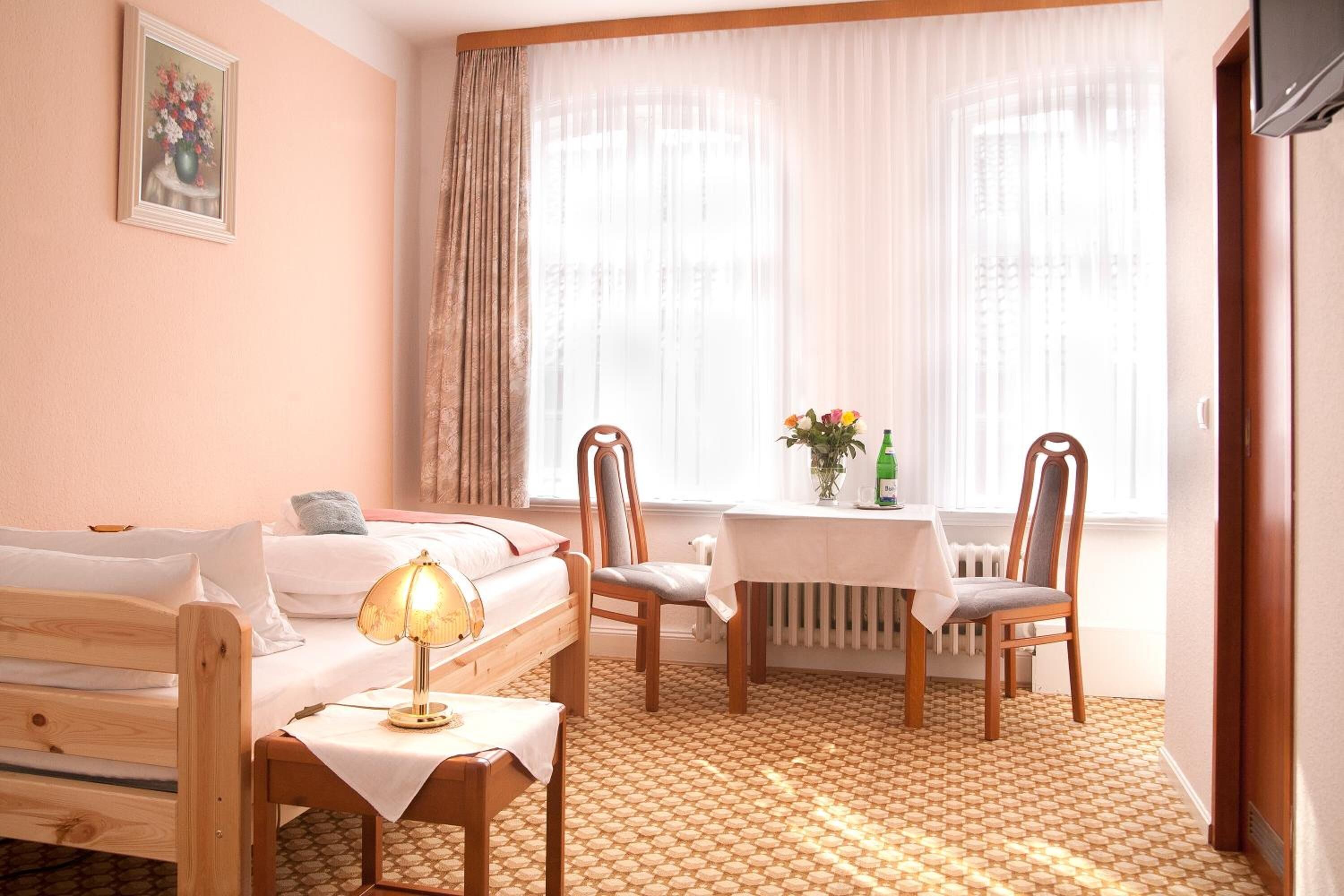 Een slaapkamer met een bed, nachtkastje, lamp, twee stoelen, een klein tafeltje met een bloevas en een raam met gordijnen.