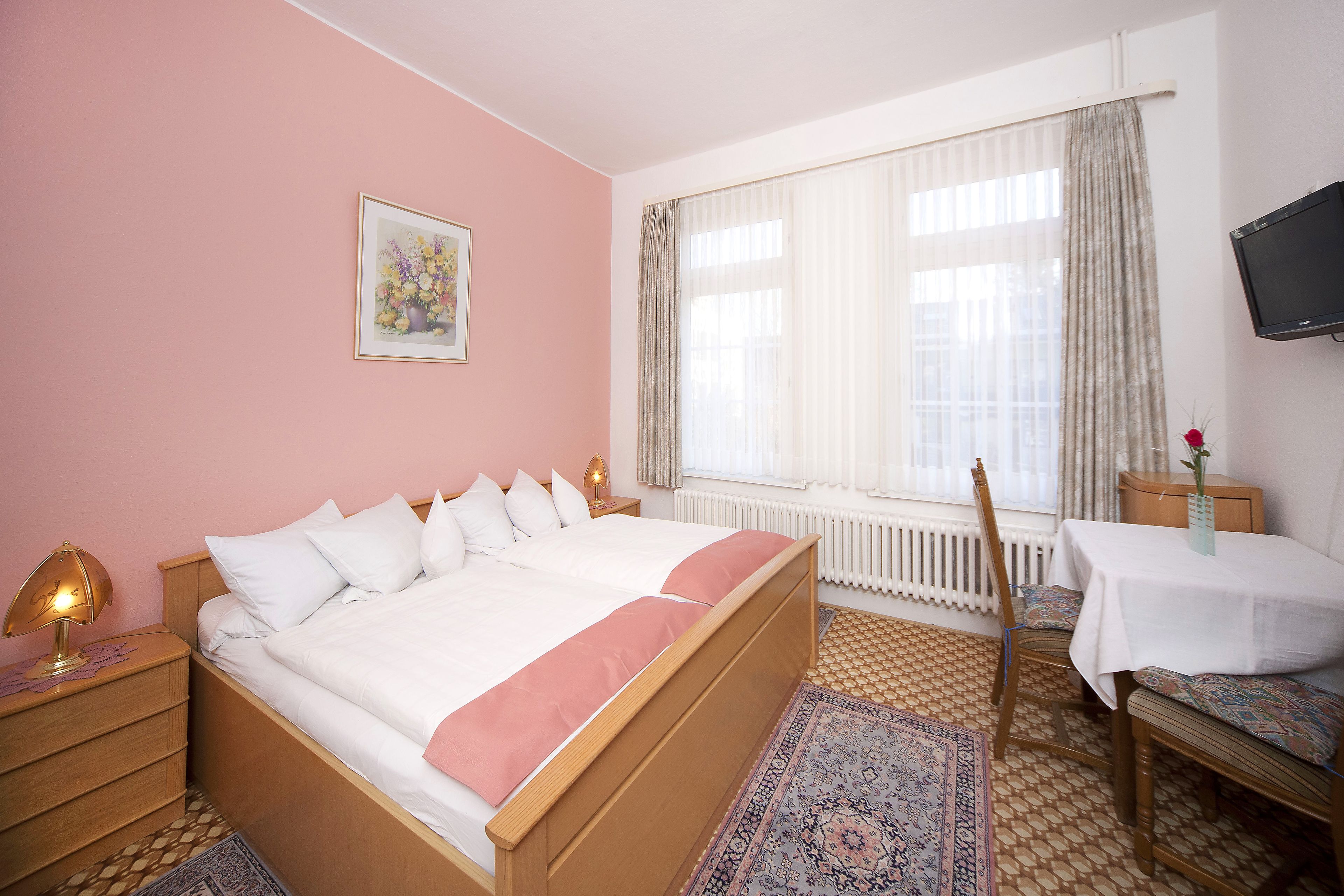 A bedroom with a bed, bedside tables, a chair, a table, a TV, and a window with curtains.