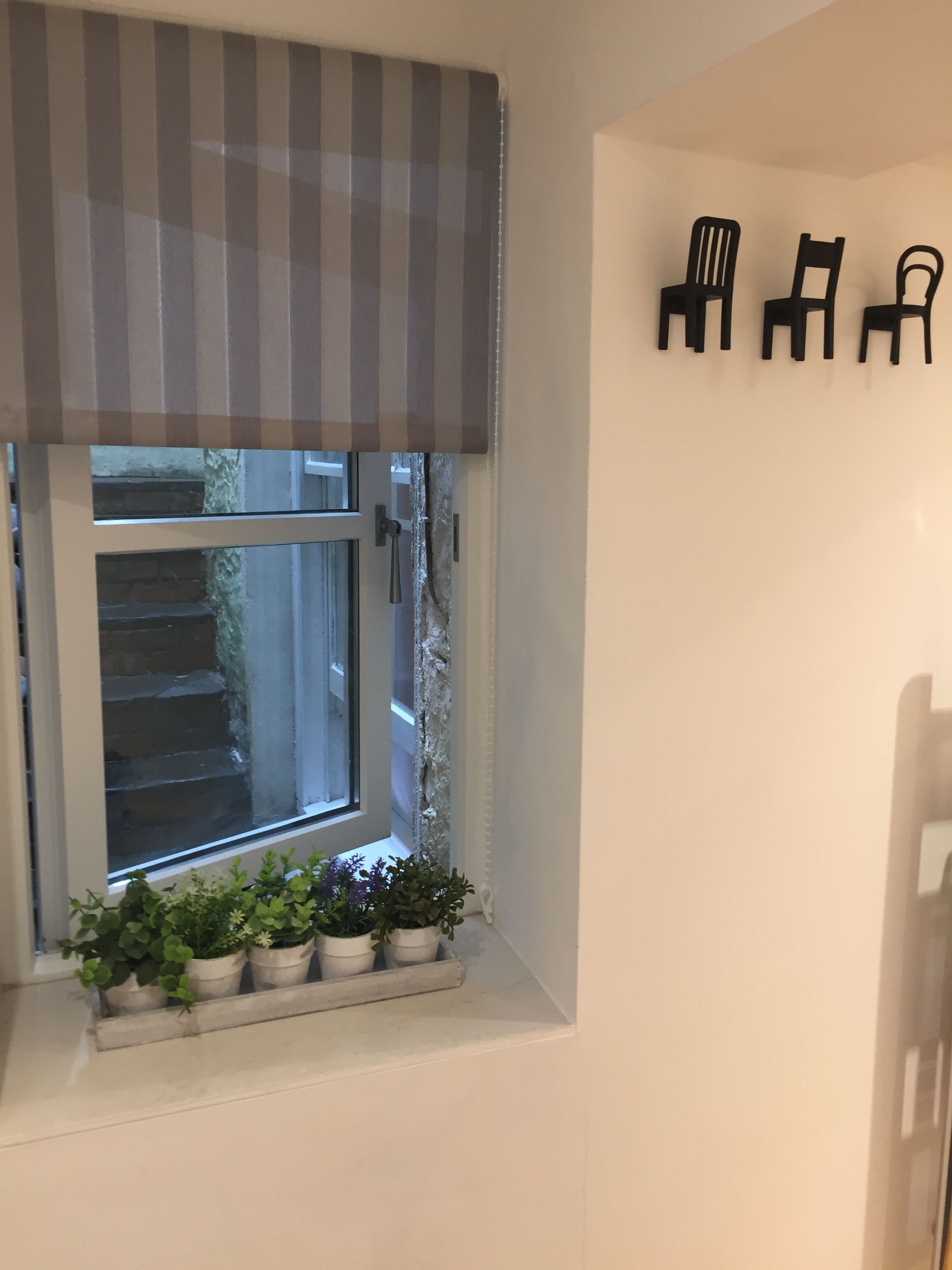A window with a striped awning, a small window sill with potted plants, and three black chairs on the wall.