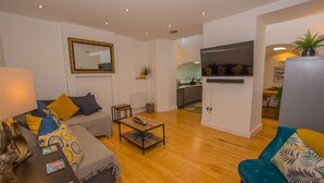 A modern living room with a sofa, armchair, and coffee table. A TV is mounted on the wall, and there is a kitchen area visible in the background. - Lancaster Apartment - Central Tenby (Tenby)