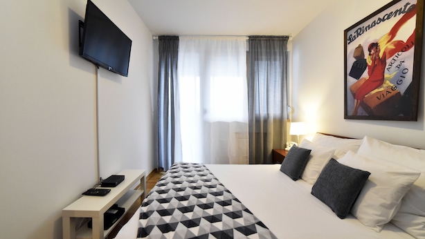 A hotel room with a bed, a TV mounted on the wall, a desk with a lamp, and a window with curtains. - Botanic Apartment (Zagreb)