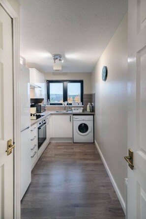 A narrow kitchen with a washing machine, a window, and a clock on the wall.