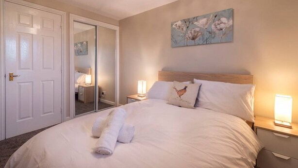 Un lit bien fait, avec du linge de lit blanc et une serviette enroulée, une tête de lit en bois, une table de chevet avec une lampe et un tableau de fleurs accroché au mur.