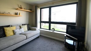 Un salon comprenant un canapé, un téléviseur, une fenêtre donnant sur un beau paysage et une étagère ornée d’objets décoratifs.