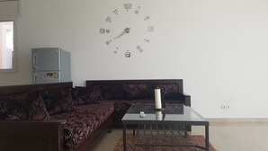 A living room with a dark patterned sofa, a glass coffee table, and a wall clock. - Dar Lebharr (Kelibia)