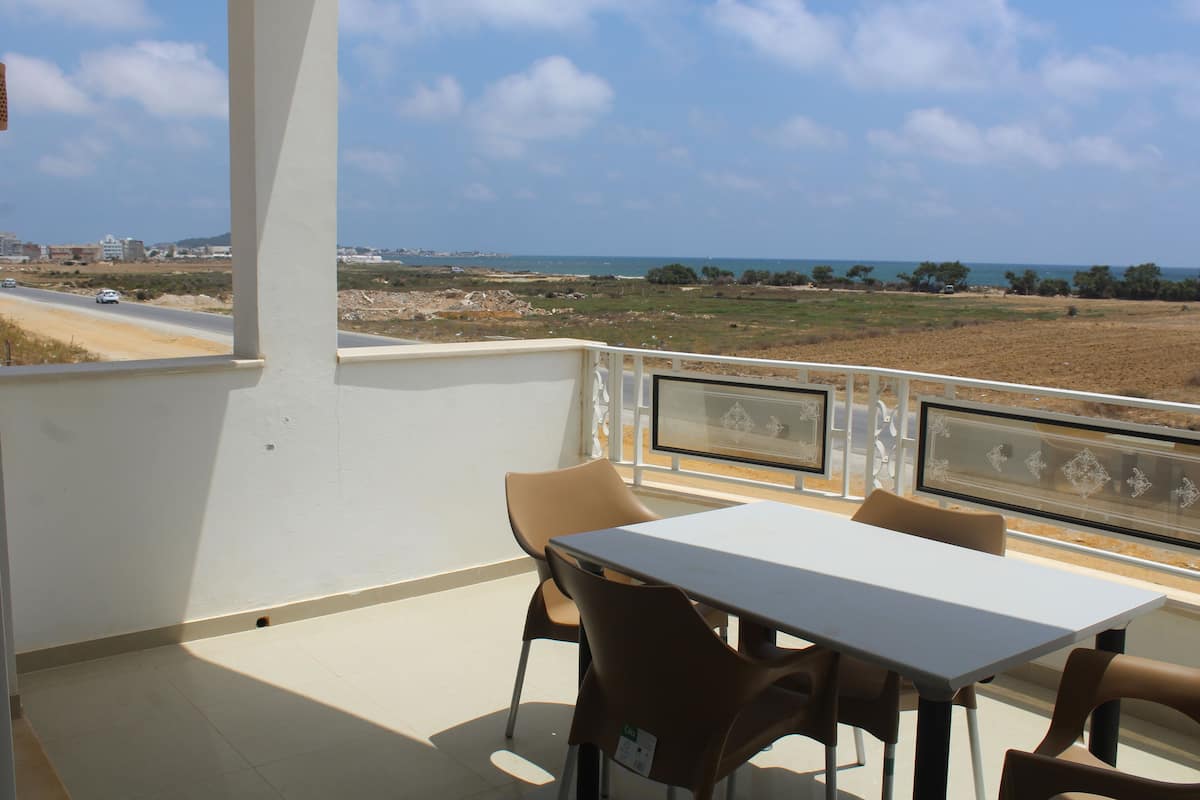 A balcony with a table and chairs, overlooking a coastal landscape with buildings and a road.