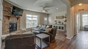 A spacious living room with a fireplace, a flat-screen TV mounted on a brick wall, a ceiling fan, and hardwood flooring.