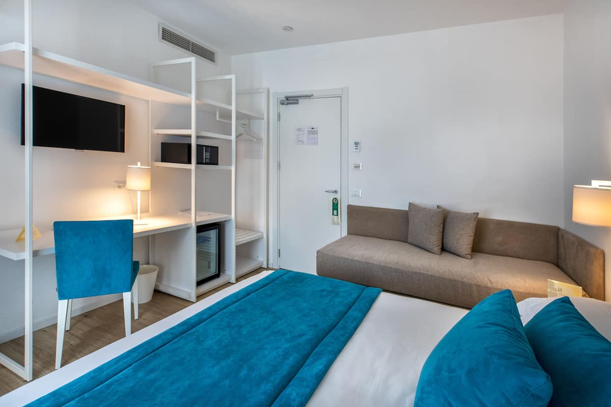 A hotel room with a bed, a desk with a chair, a television, a sofa, and a door.