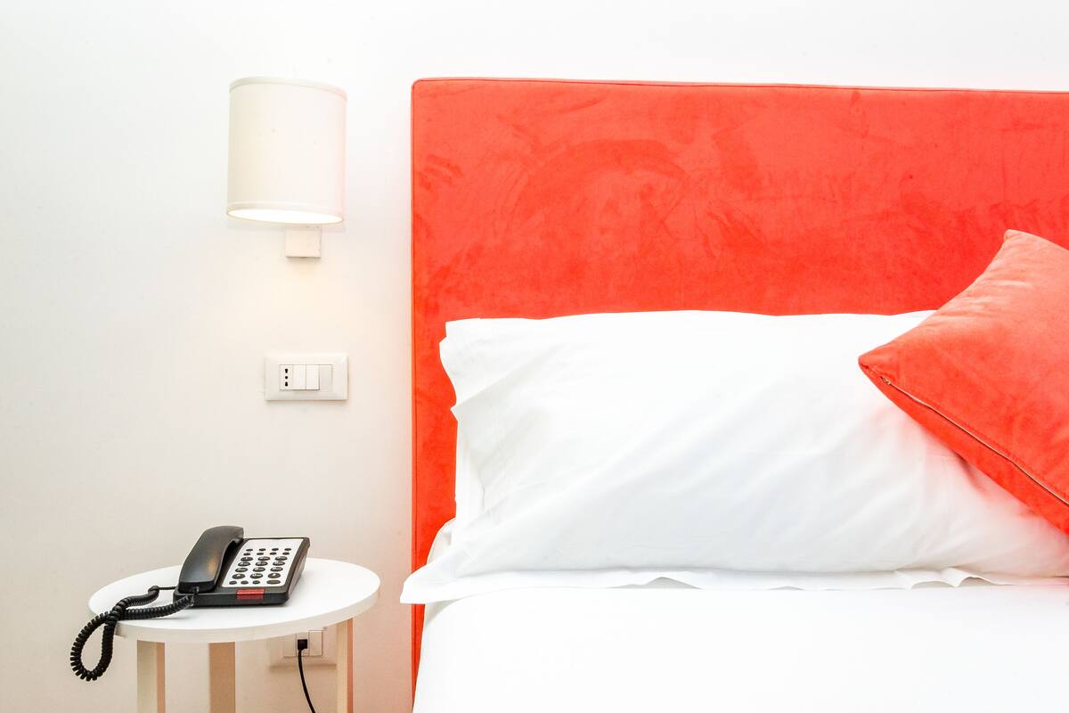 A hotel room with a bed, a bedside table with a phone, and a wall-mounted light.