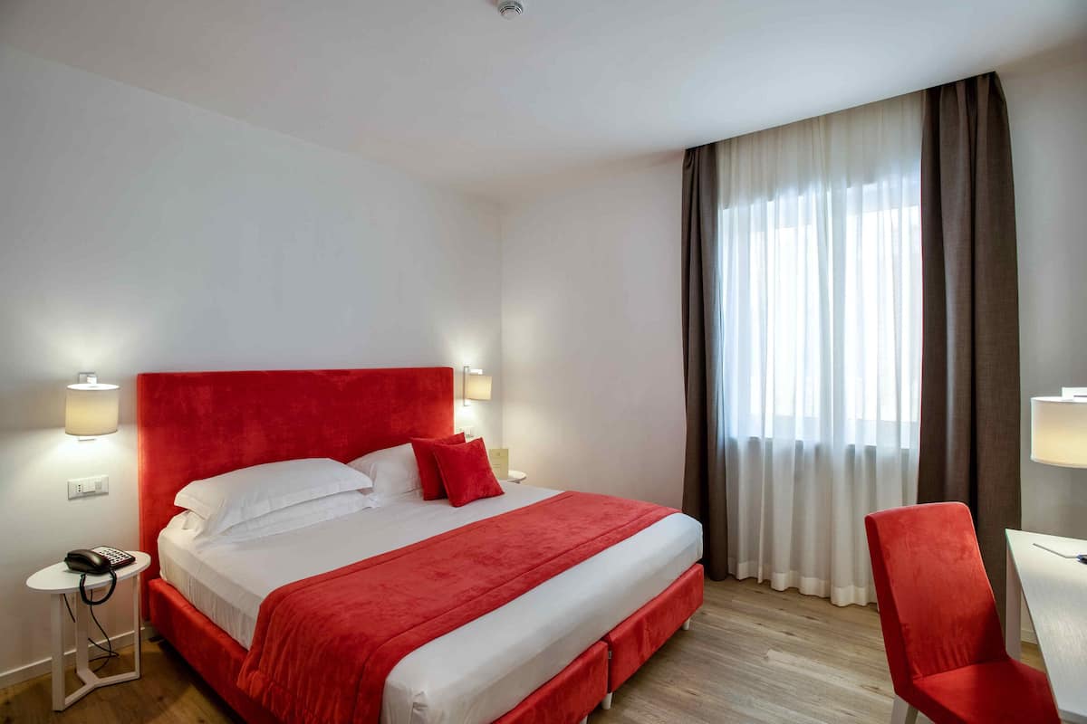 A hotel room with a large bed, a red headboard, a bedside table with a lamp, a chair, and a window with curtains.