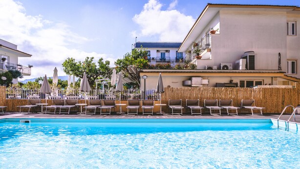A swimming pool with lounge chairs and umbrellas, surrounded by buildings with balconies and outdoor furniture. - Hotel Isabella (Sant'Agnello)