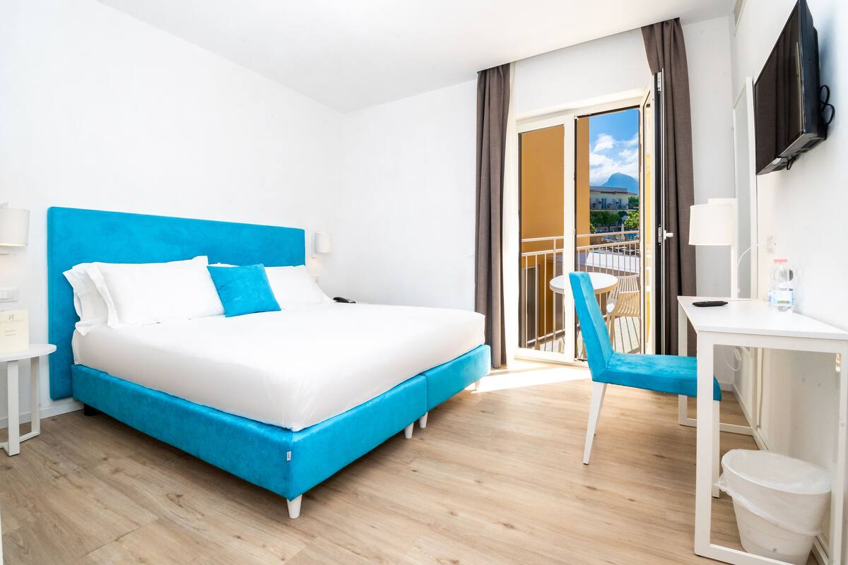 A hotel room with a bed, a desk, a chair, and a balcony with a view.