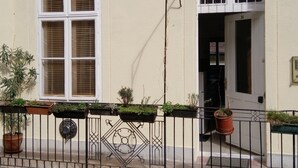 A building with a white facade, a large window with blinds, and a door with a small window above it. There are potted plants on the ground floor and a metal fence with decorative elements. - Classical Budapest apartment in the old Pest (Budapest)