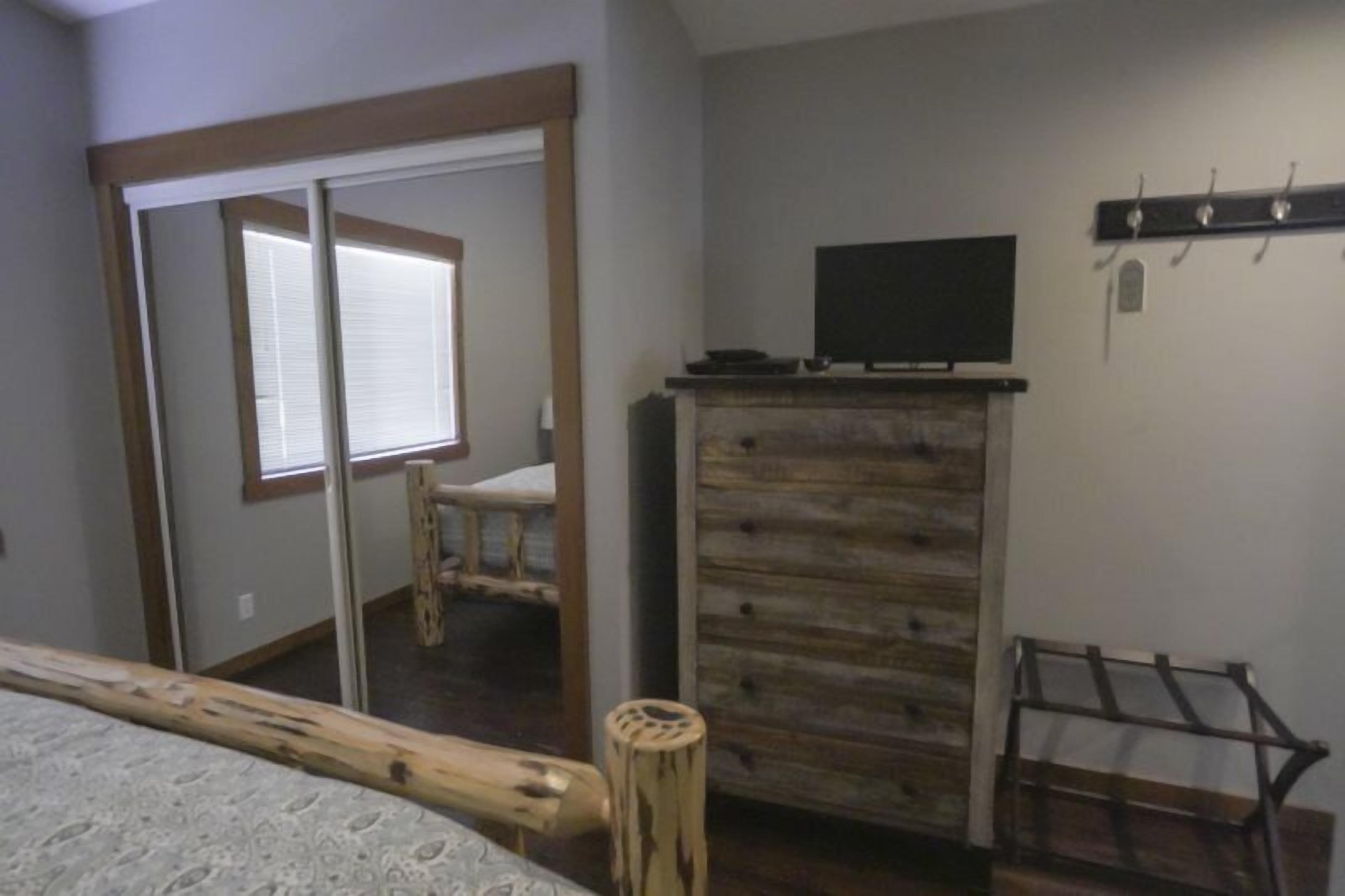 A bedroom with a bed, a wooden dresser, a television on top, and a window with blinds.