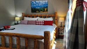 Een slaapkamer met een houten hoofdbord, een bed met wit beddengoed, rode en geruite kussens, een nachtkastje met een lamp, en een schilderij van een berglandschap aan de muur.