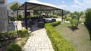 An outdoor patio area with stone flooring, covered seating, and a garden with hedges and palm trees. - Serravalle Relais & Country Villa (Chiaramonte Gulfi)