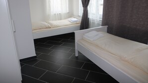 Ein Schlafzimmer mit zwei Einzelbetten, einem Fenster mit durchsichtigen Vorhängen, einem dunkel gefliesten Boden und einem weißen Kleiderschrank.