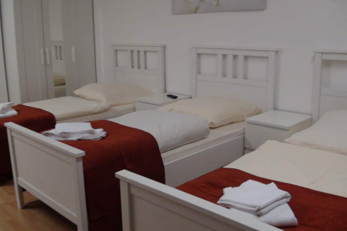 Een hotelkamer met drie enkele bedden, elk met een witte hoofdbord en een rode deken. Er staat een witte nachtkastje met een afstandsbediening erop.