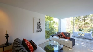 Un salon moderne avec une table basse en verre, un canapé blanc agrémenté de coussins à motifs, une œuvre d’art encadrée en noir et blanc, et une grande fenêtre donnant sur des arbres et un balcon.