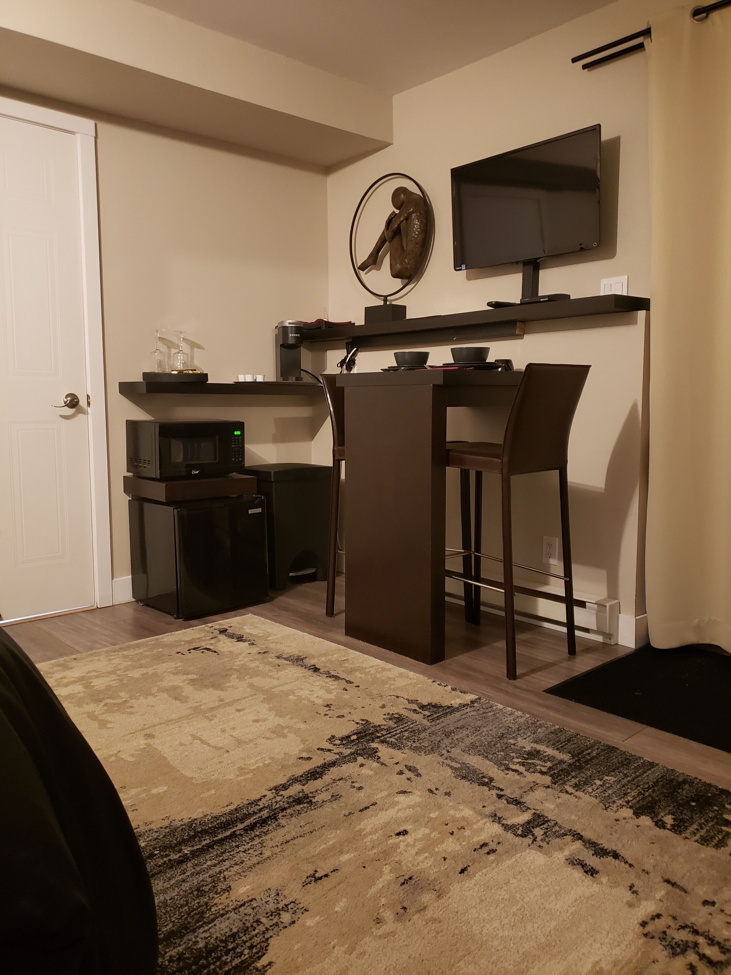 A compact living space with a kitchenette, a dining area with two chairs, a small TV mounted on the wall, and a door to the left.
