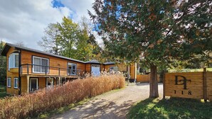 Une cabane en bois à deux étages, avec une allée en gravier, entourée de hautes herbes et d’arbres.