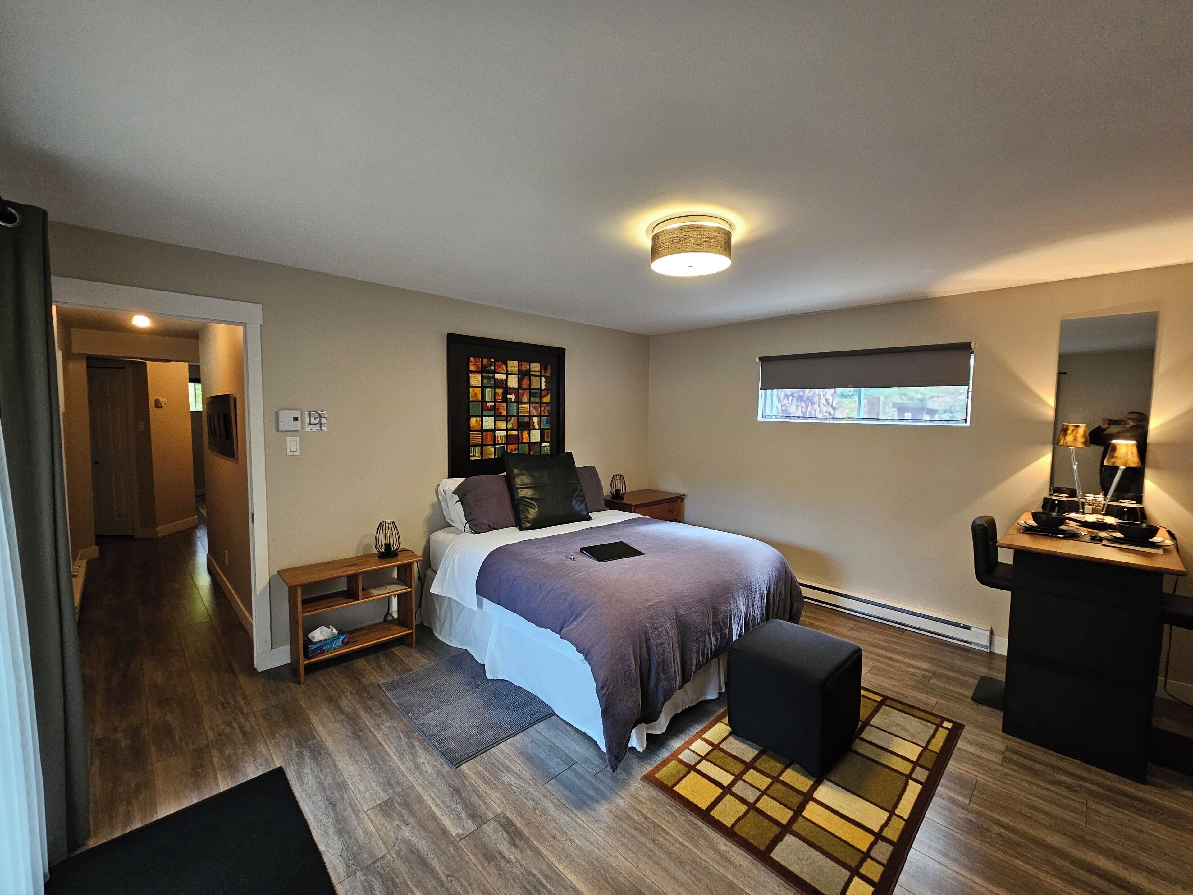 Quarto com cama, mesas de cabeceira, escrivaninha, espelho, janela e luminária no teto.