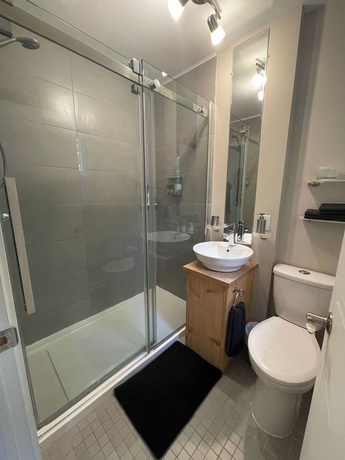 Un baño moderno con cabina de ducha de vidrio, tocador de madera con lavabo blanco y váter blanco.
