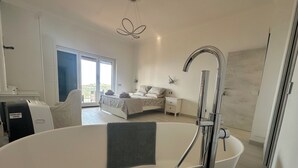A modern bedroom with a large bathtub, a bed with a patterned bedspread, a bedside table, and a view of a balcony through a large window. - Villa Asia (Massa Lubrense)
