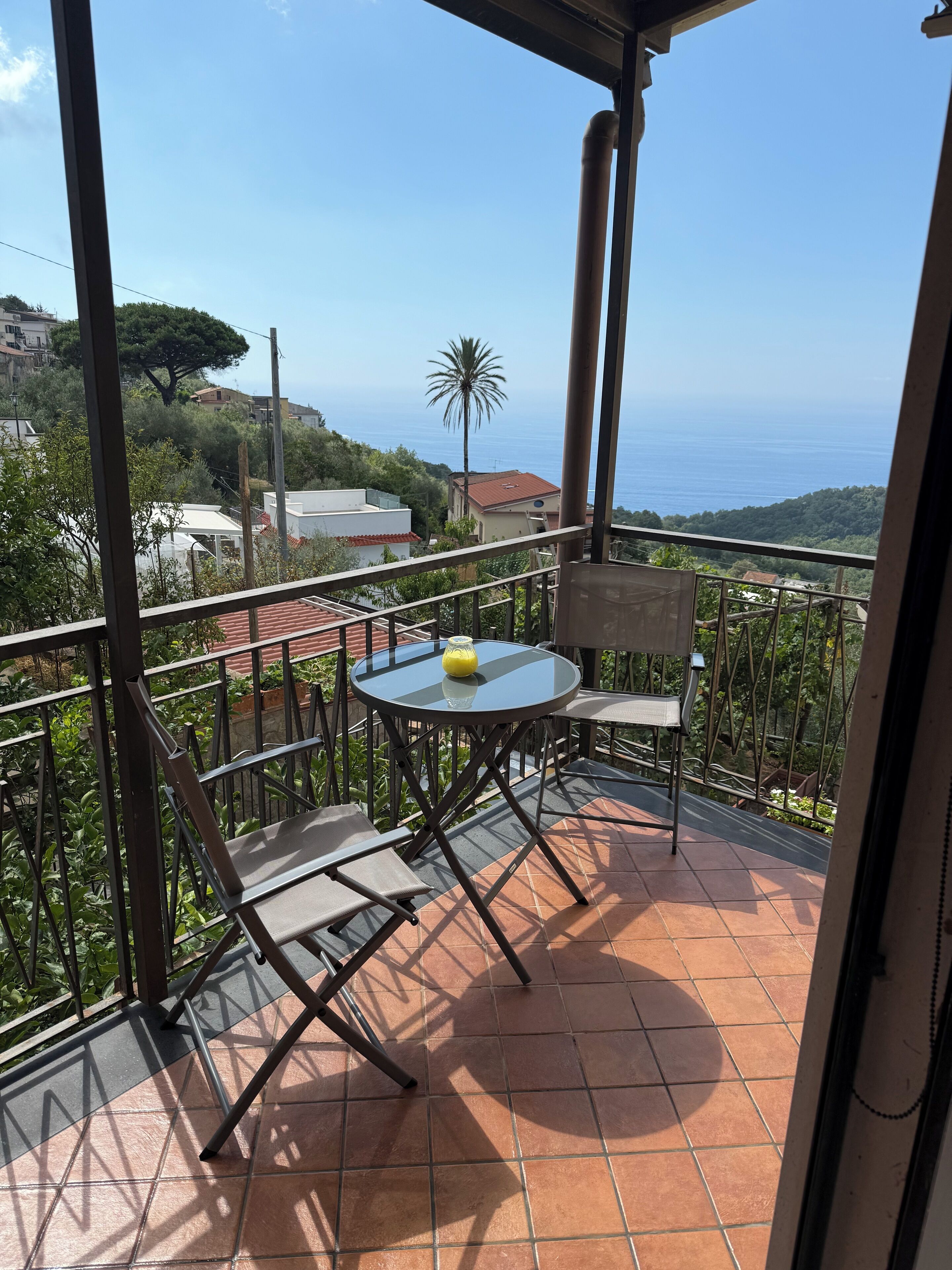 A balcony with a table and chairs, overlooking a coastal landscape with houses and a palm tree.