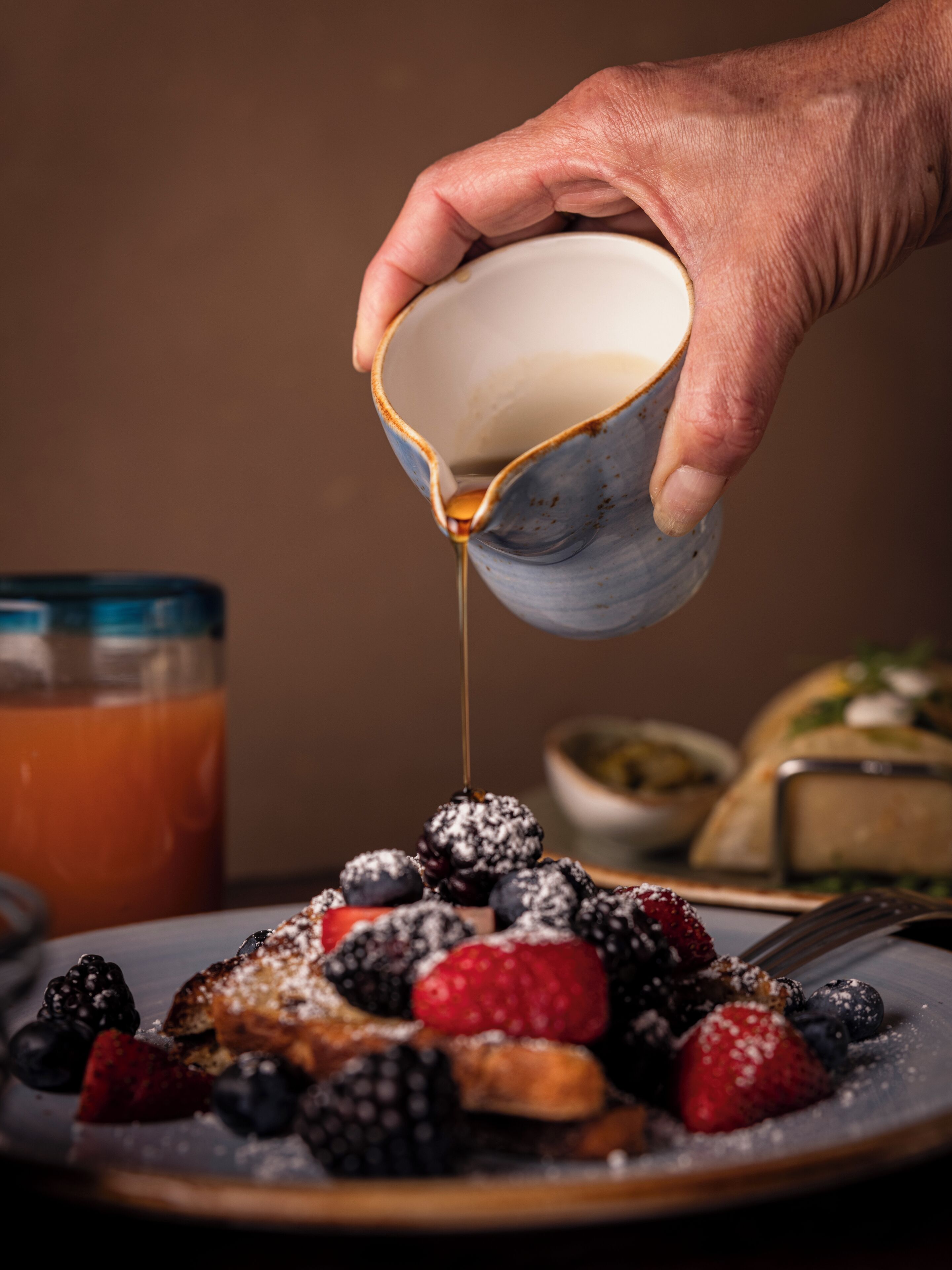 Une main verse du sirop sur une assiette de crêpes garnies de fruits des bois.
