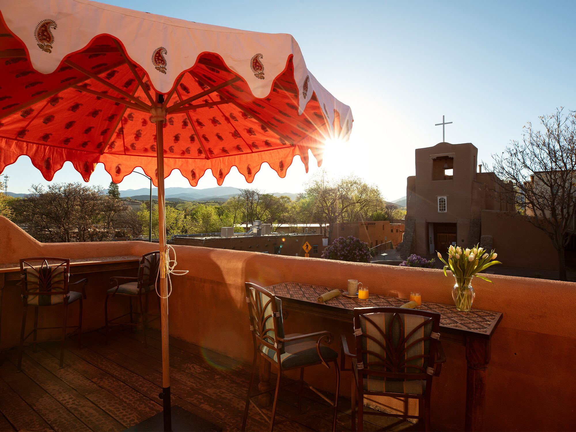 An outdoor patio with wooden furniture, a table with a vase of flowers, and a view of a church and mountains in the background.