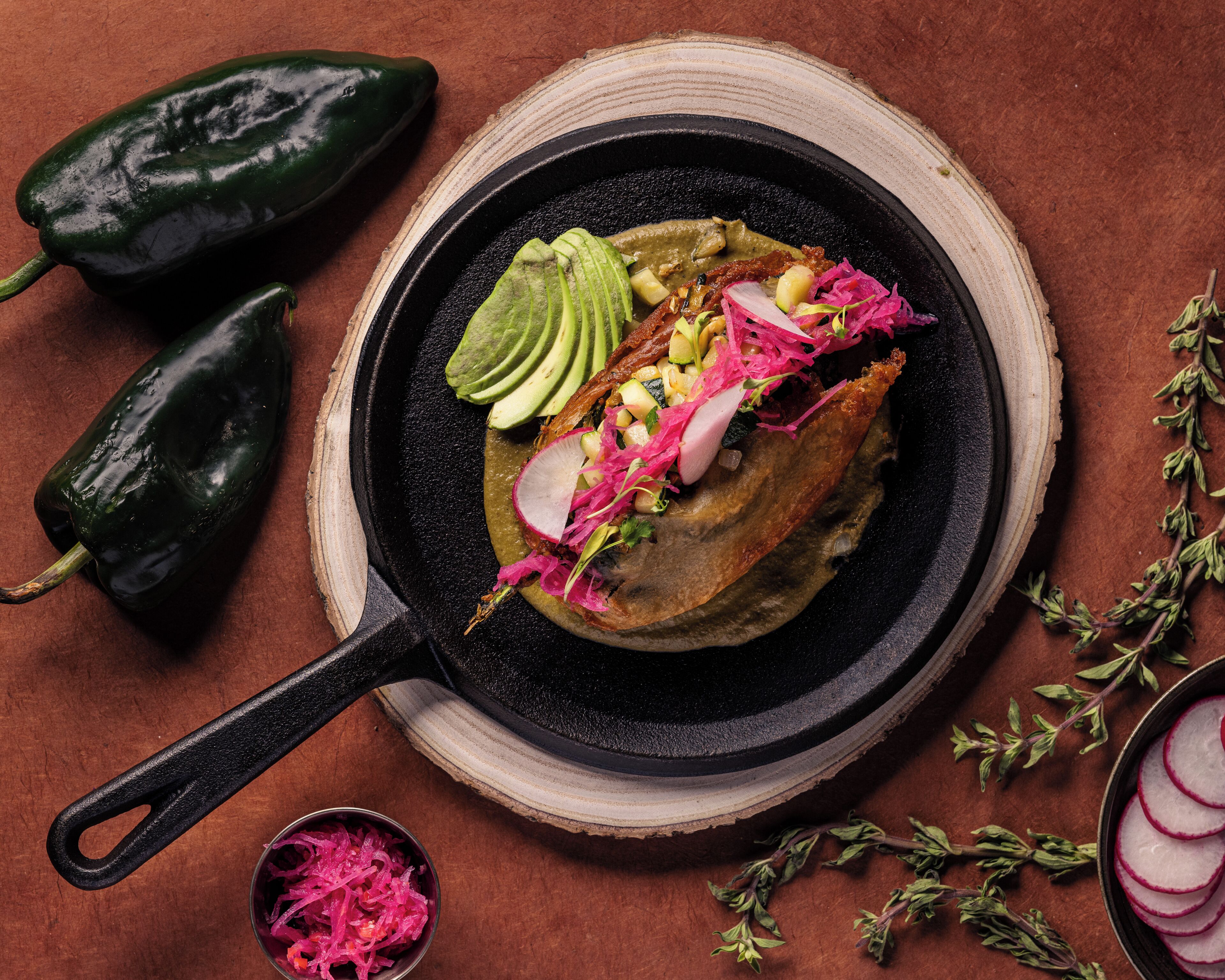 Eine Pfanne mit einer Speise aus Avocado-Scheiben, Rettich und anderem Gemüse, umgeben von ganzen Avocados und Kräutern auf einer Holzbrett.