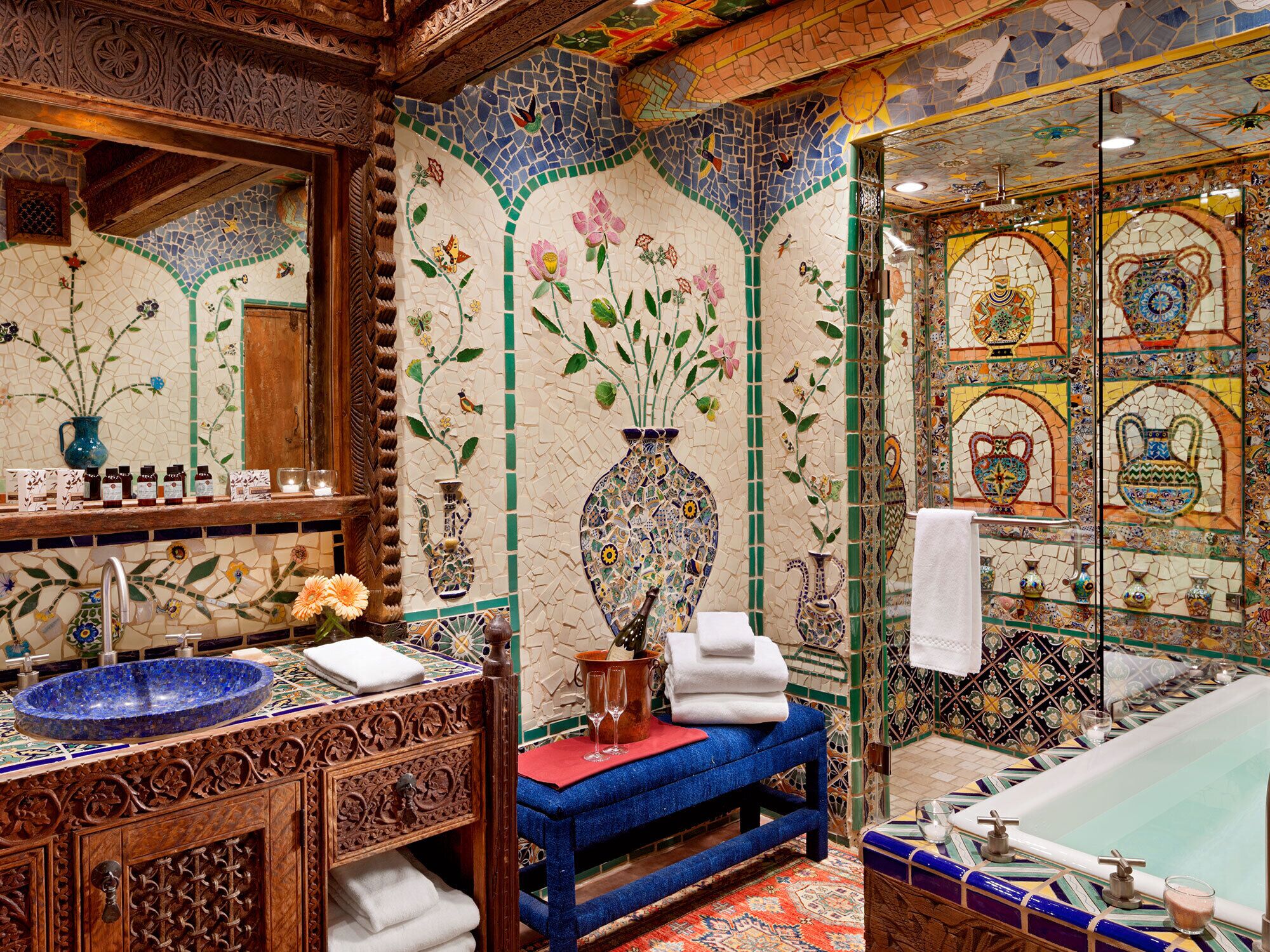 Une salle de bain avec un mur décoré de mosaïques, un banc bleu, un lavabo avec un receveur bleu et une baignoire dont les parois sont recouvertes de carreaux.