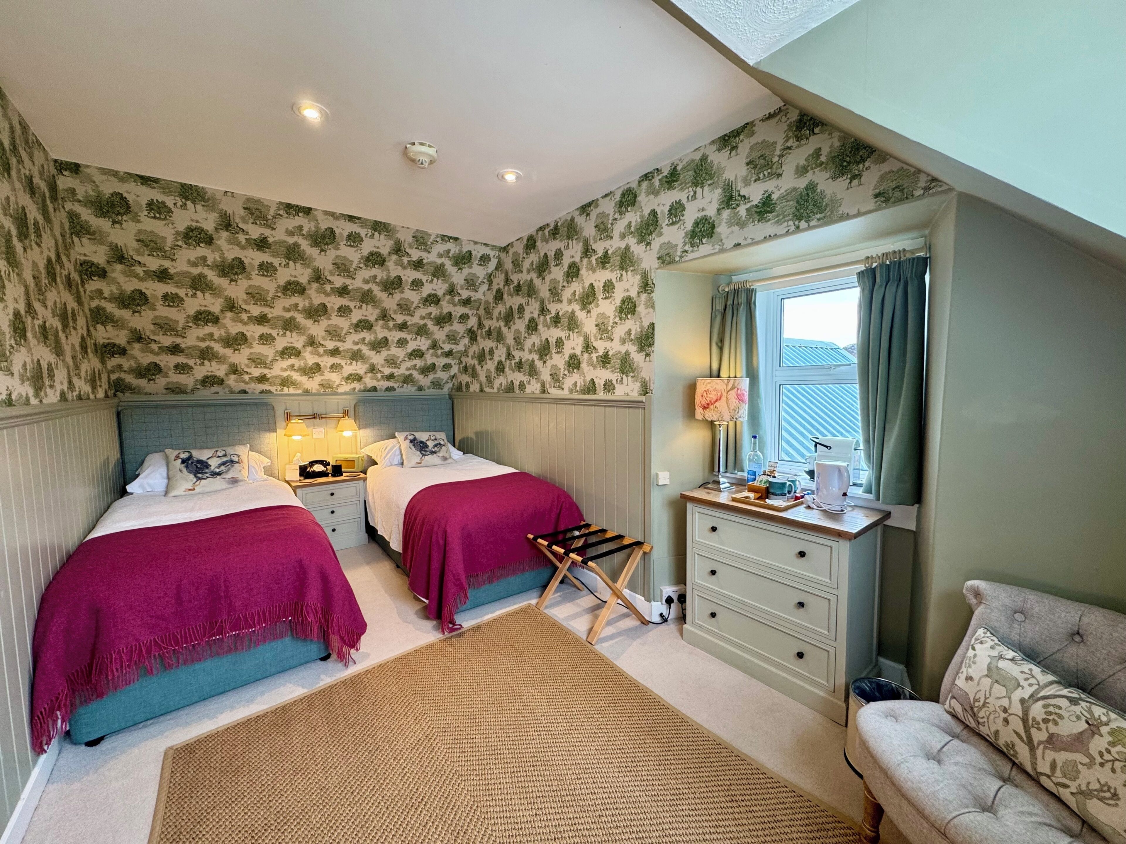 A bedroom with two beds, a desk, a chair, a window with curtains, and a patterned wallpaper.